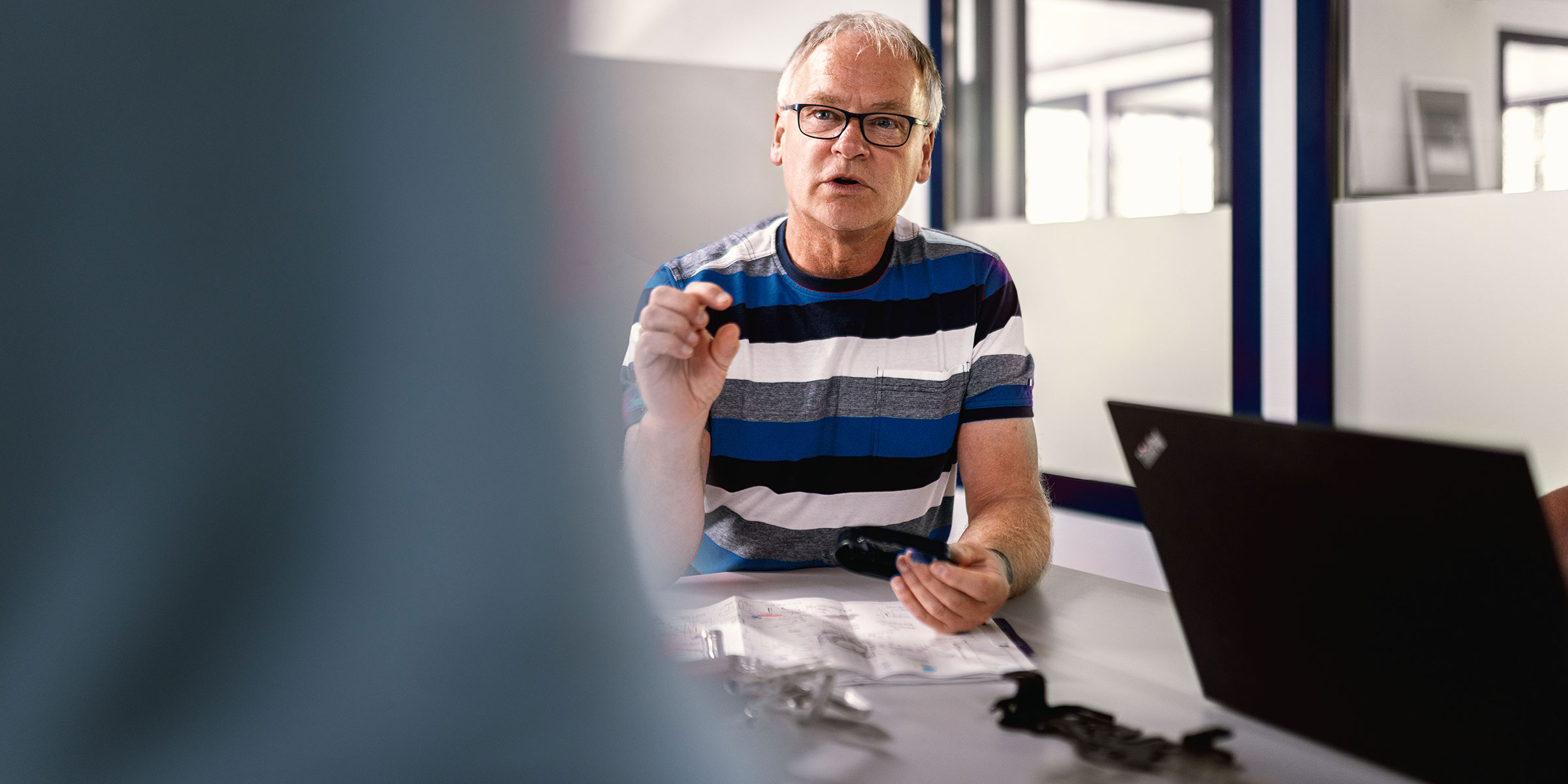 Ein Mitarbeiter erklärt an einem Stehtisch mit Laptop und Bauteilen den aktuellen Projektstand seinen Kollegen.