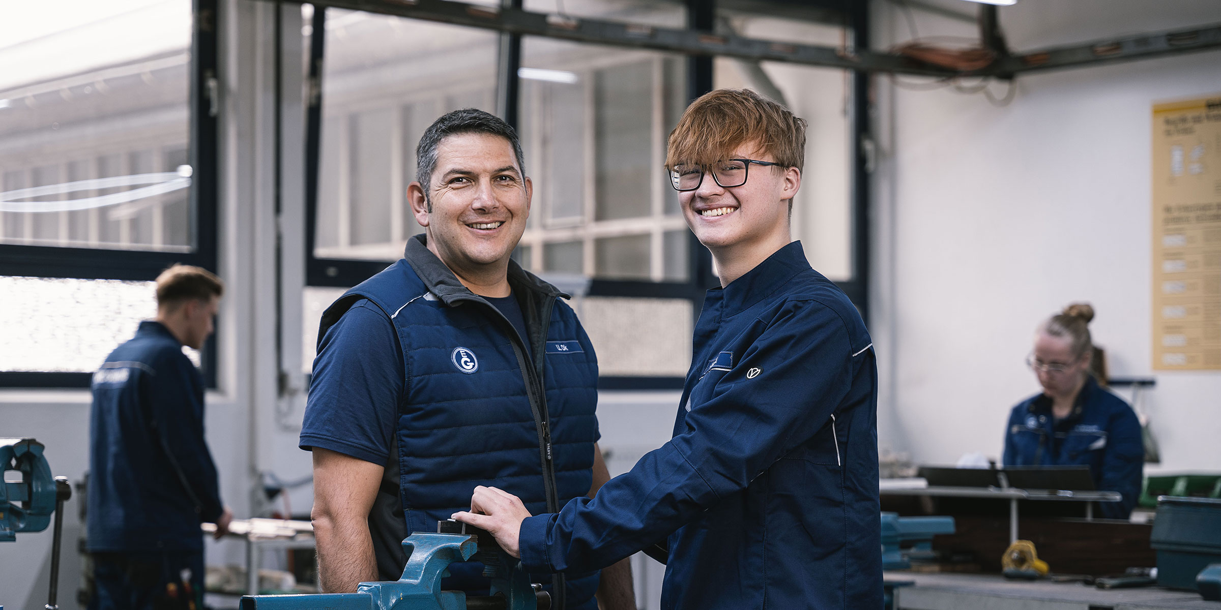 Ausbilder und Auszubildender arbeiten gemeinsam in einer Werkstatt der Stanztechnik. Beide tragen Arbeitskleidung und stehen lächelnd an einer Werkbank, während im Hintergrund weitere Auszubildende praktisch arbeiten.